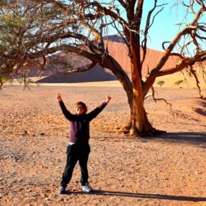 Enfant bras levés devant un arbre mort, au pied des dunes rouges du désert du Namib